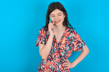 Portrait of successful joyful young caucasian woman wearing floral dress over blue background talking on mobile phone with friend. Lifestyle and communication concept