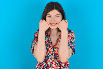 Happy young caucasian woman wearing floral dress over blue background keeps fists on cheeks smiles broadly and has positive expression being in good mood