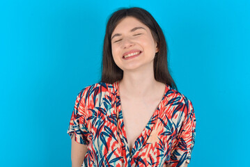 Positive young caucasian woman wearing floral dress over blue background with overjoyed expression closes eyes and laughs shows white perfect teeth