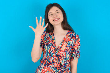 Fototapeta premium young caucasian woman wearing floral dress over blue background smiling and looking friendly, showing number five or fifth with hand forward, counting down