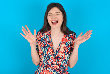 Crazy outraged young caucasian woman wearing floral dress over blue background screams loudly and gestures angrily yells furiously. Negative human emotions feelings concept