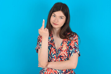 young caucasian woman wearing floral dress over blue background shows middle finger bad sign asks not to bother. Provocation and rude attitude.