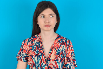 Dissatisfied young caucasian woman wearing floral dress over blue background purses lips and has unhappy expression looks away stands offended. Depressed frustrated model.