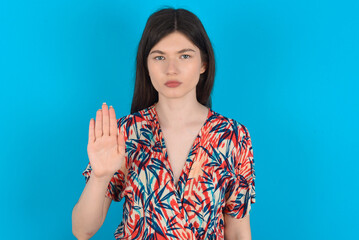 young caucasian woman wearing floral dress over blue background shows stop sign prohibition symbol keeps palm forward to camera with strict expression