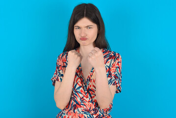 Displeased annoyed young caucasian woman wearing floral dress over blue wall clenches fists, gestures pissed, ready to revenge, looks with aggression at camera stands full of hate, being pressured