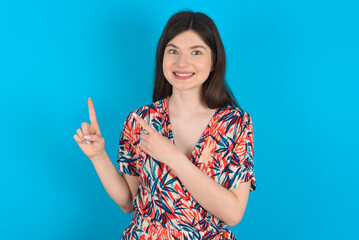 young caucasian woman wearing floral dress over blue background points at copy space indicates for advertising gives right direction