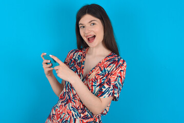 Nice addicted cheerful young caucasian woman wearing floral dress over blue background using gadget playing network game
