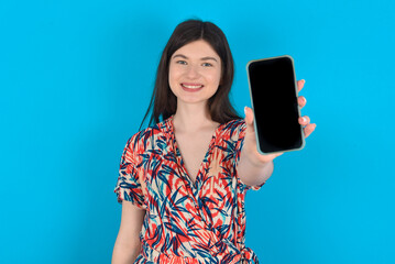 Naklejka premium Charming adorable young caucasian woman wearing floral dress over blue background holding modern device, showing black screen smartphone