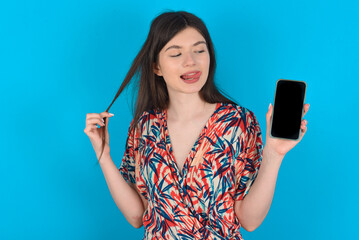 Photo of nice pretty young caucasian woman wearing floral dress over blue background demonstrate phone screen hold hair tails