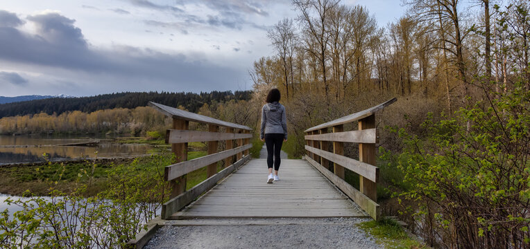 Woman Walking on a Wooden Path with green trees in Shoreline Trail, Port Moody, Greater Vancouver, British Columbia, Canada. Trail in a Modern City during a Cloudy Sunset.