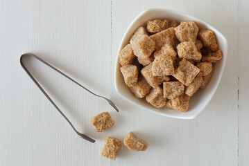 Cube-shaped cane sugar in wooden bowl on a white wooden table with silver sugar tongs.  Flat lay, top down view, no people.