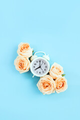 White alarm clock surrounded by cream roses on blue background. Florescence time concept. Vertical banner, copy space