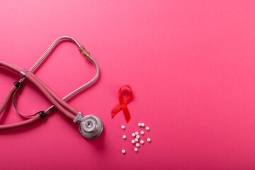 Directly above shot of red aids awareness ribbon with stethoscope and medicines on pink background