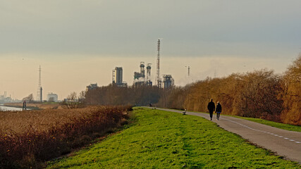 People walking on a pth on the dam along river Scheldt in Antwerp, Flanders, Belgiumwith petrochemical industrial infrastructure in the distance 