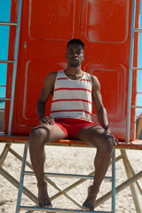 Obraz na płótnie Canvas Portrait of confident african american young man sitting on lifeguard hut at beach on sunny day