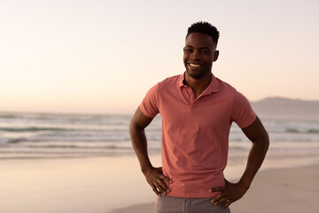 Portrait of smiling african american young man with arms akimbo standing at beach against clear sky