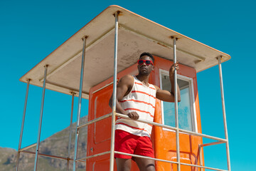 African american young man wearing sunglasses standing on lifeguard hut against clear blue sky