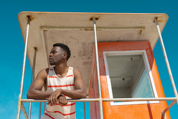 Low angle view of african american young man looking away standing on lifeguard hut against blue sky