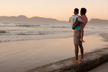 African american young man carrying son while standing on rock at beach against clear sky at sunset