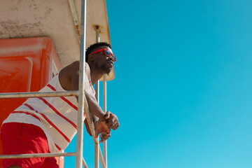 Low angle view of african american young man in sunglasses standing on lifeguard hut against sky