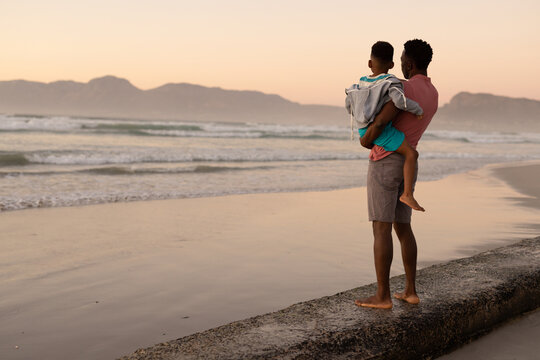 African American Young Man Carrying Son While Standing On Rock At Beach Against Clear Sky At Sunset