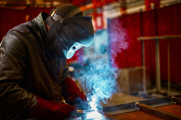 Skilled worker in protective mask welds metal part at plant