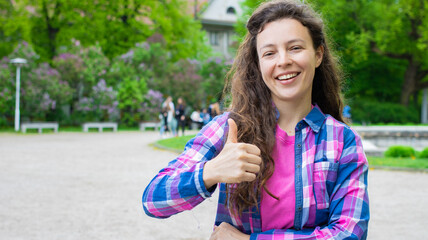 Smiling young pretty woman student in pink blue cloth showing thumb up approval like gesture, support satisfied recommend the best, standing in green city park outdoors. Good job sign. Copy space