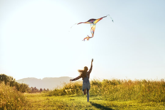 Long-haired Girl with flying a colorful kite on the high grass meadow in the mountain fields. Happy childhood moments or outdoor time spending concept image.