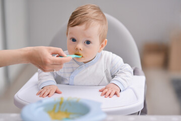 Cute baby boy sitting on a white high chair. Mother giving healthy food by spoon in the morning at home