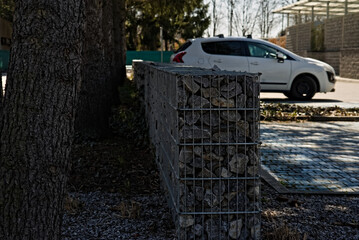 Mały murek - płotek  ze stalowej siatki i kamieni . Na parkingu wśród drzew .