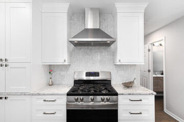 A stainless steel oven and hood in a white kitchen with marble countertops, a herringbone...