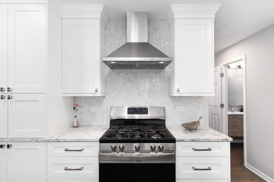A stainless steel oven and hood in a white kitchen with marble countertops, a herringbone backsplash, and a bathroom down the hall. 