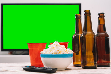 A few bottles of beer with popcorn on a table in front of a green screen TV. sporting event, party, watching a movie.
