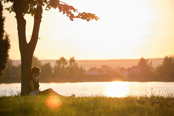Lonely woman sitting alone on green grass lawn leaning to tree trunk on lake shore on warm evening. Solitude and relaxing in nature concept - Powered by Adobe