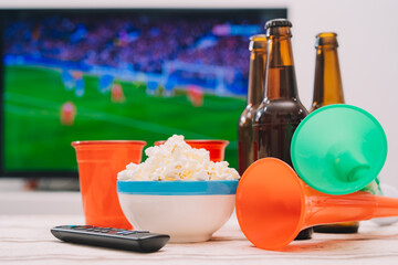 A beer with popcorn and trumpets to cheer on his football team, in front of the TV screen showing a sporting event.