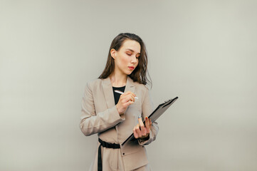 Naklejka premium Attractive woman in a suit stands on a beige background with a tablet in hand and writes with a pen on paper with a serious face. Businesswoman makes notes.