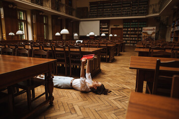 A female student lies on the ground of a public old library and reads a book.