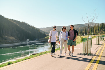 A positive group of friends of tourists in casual clothes walk around the mountain resort in summer and communicate with smiles on their faces.