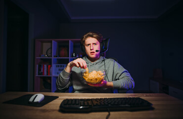 Serious young man in a headset sits at night at the computer and eats chips from a plate, looking intently at the camera.
