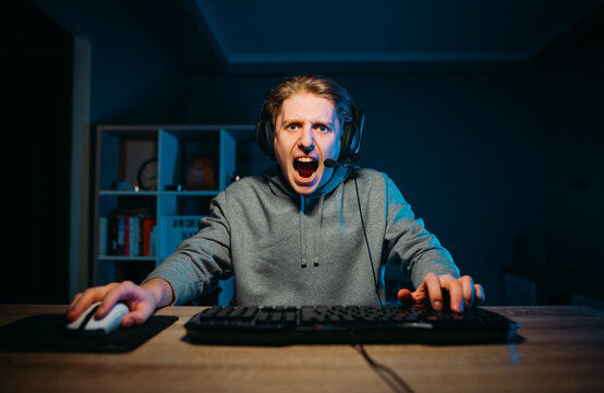 Angry guy in a headset plays online games on the computer at night, looks at the camera and shouts.