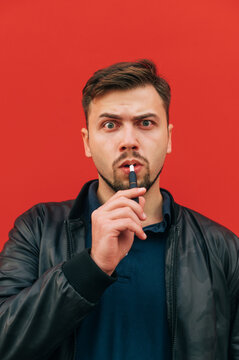 Portrait of a funny man in dark clothes smokes an electronic cigarette on a red background and looks at the camera. Vertical