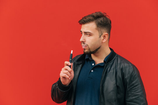 Adult handsome man in dark clothes stands against the backdrop of a red wall and smokes iqos dilvlaich aside