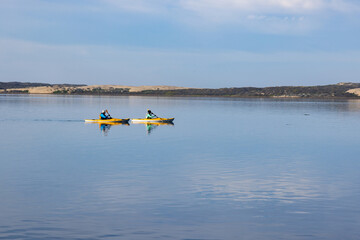 Two yellow Kayaks glide through calm water with clouds reflecting in the water, with sand dunes in the background. Morro Bay, California