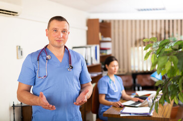 Friendly doctor wearing blue uniform with phonendoscope on his neck welcoming to medical office, making inviting gesture with hands