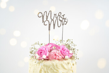 Wedding 2 Tiered Cake with Mr and Mrs topper. Styled with fresh pink roses and gypsophila flowers, against a white background with bokeh party fairy lights. Close up. - Powered by Adobe