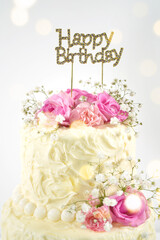 Happy Birthday 2 Tiered Cake with Happy Birthday topper. Styled with fresh pink roses and gypsophila flowers, against a white background with bokeh party fairy lights. Vertical orientation.