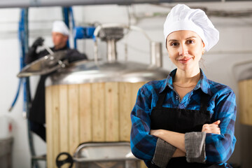 Portrait of confident smiling young female brewer standing with crossed arms in craft brewery...