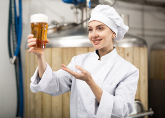 Portrait of cheerful female brewmaster in white uniform presenting glass of fresh beer.