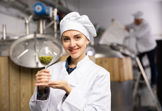 Portrait of positive woman brewmaster holding glass of hops pellets in beer factory. - Powered by Adobe