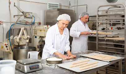 Experienced middle aged woman working with male assistant in bakery, forming baguettes from raw dough ..